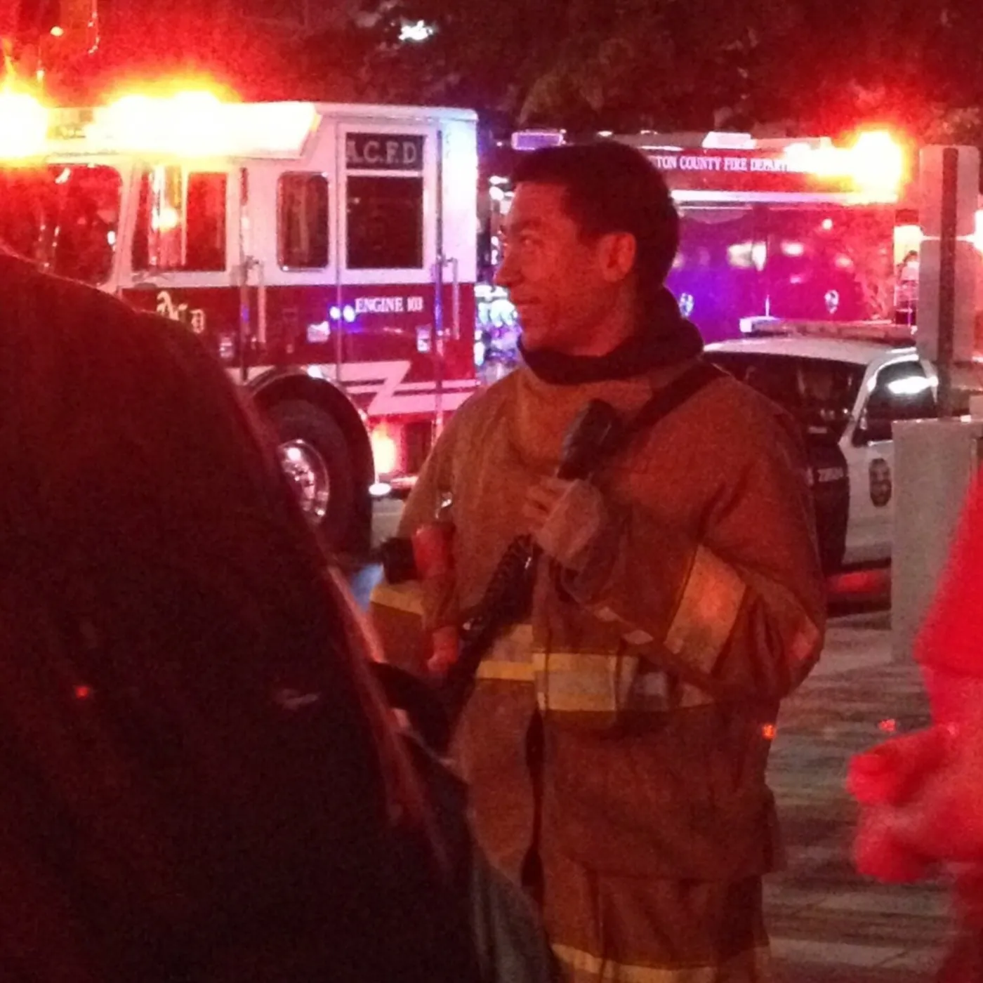 A firefighter in turnout gear at night, beside fire engines with their lights on