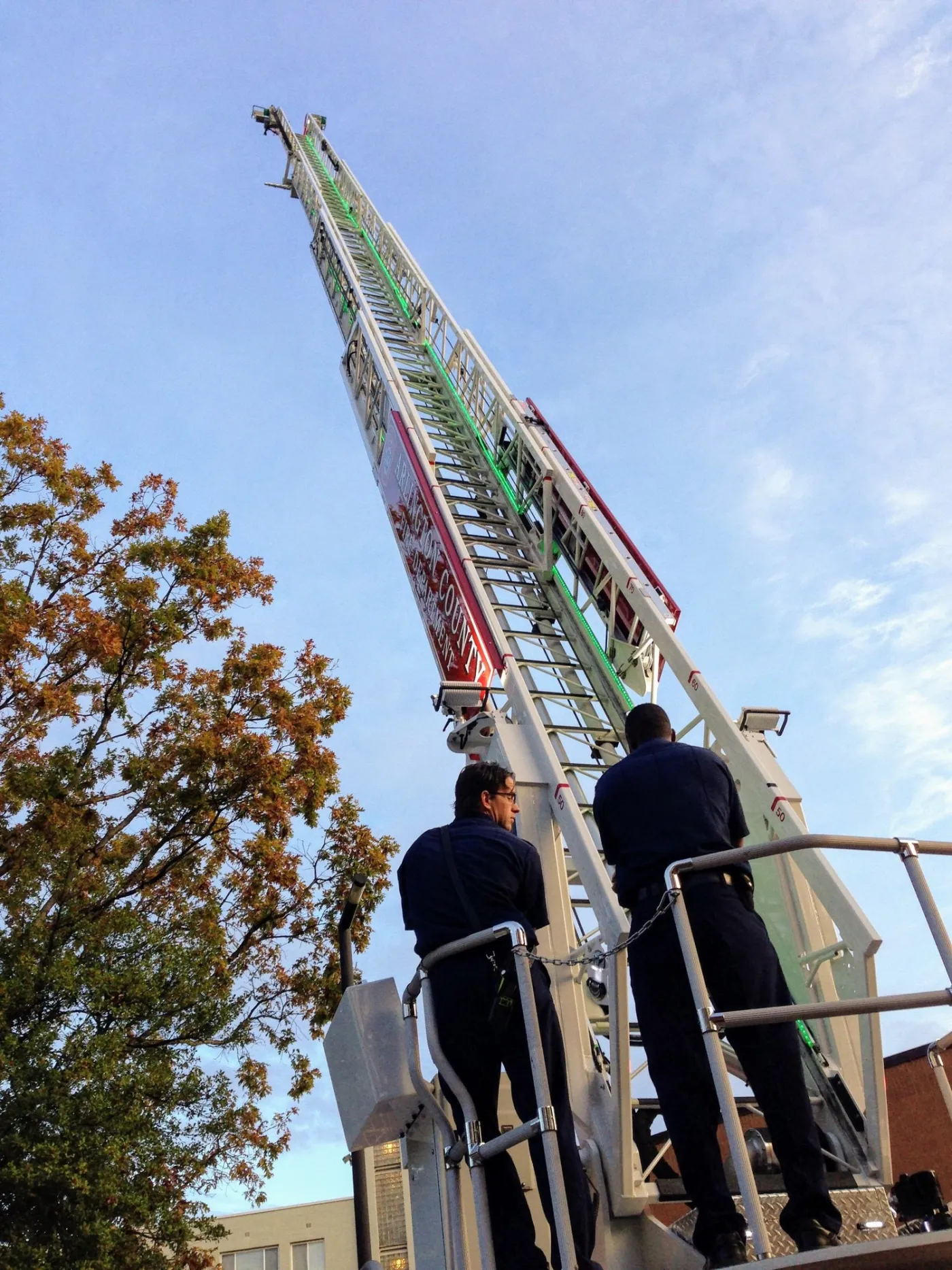 Two firefighters at the base of a tall aerial ladder raised into the sky